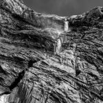 A stunning black and white capture of a waterfall flowing down rugged rock formations in a mountainous landscape.